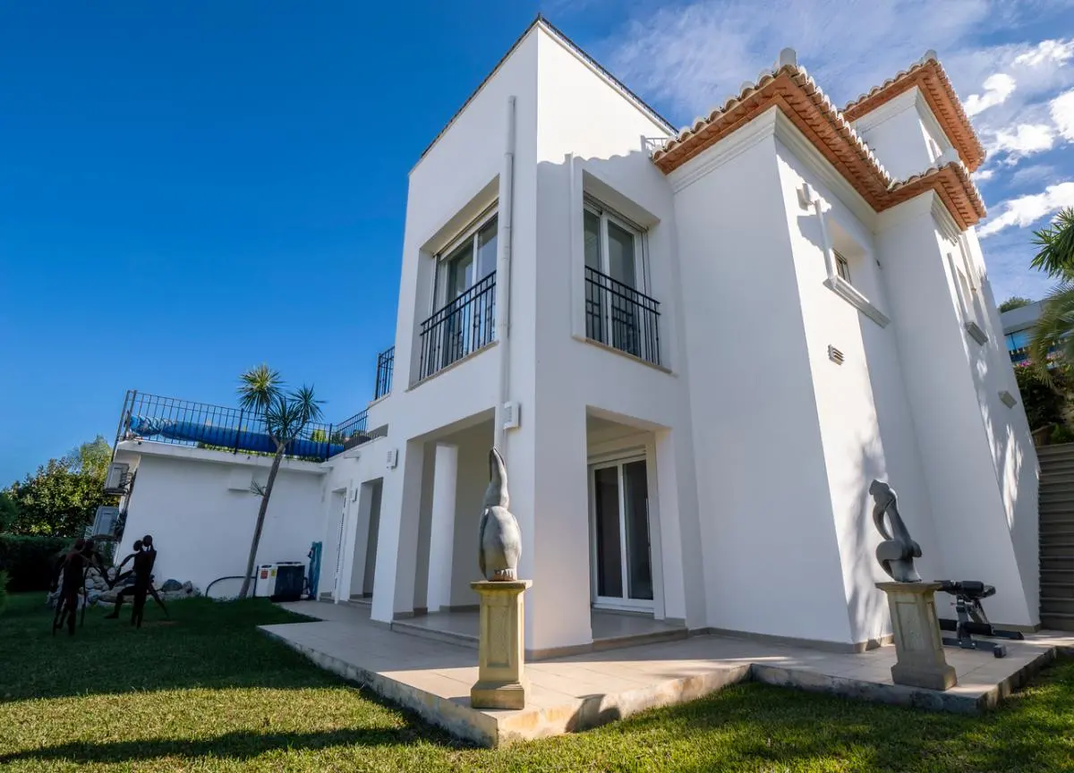 Vue extérieure d'une villa blanche à plusieurs niveaux avec toiture en tuiles, garde-corps en fer noir et jardin paysager orné de sculptures sur socles.