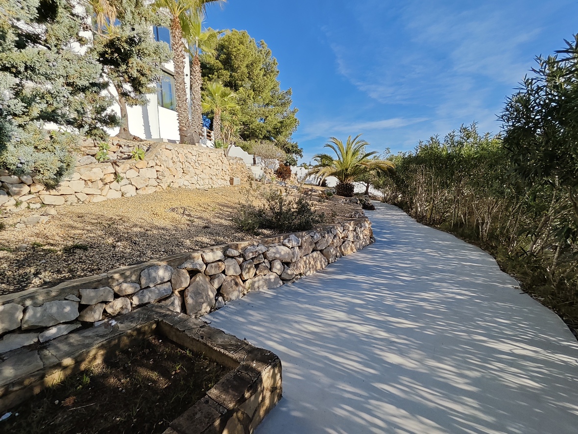 Allée pavée sinueuse traversant un jardin méditerranéen avec des murets en pierre sèche, des palmiers et des pins bordant la façade de la villa.