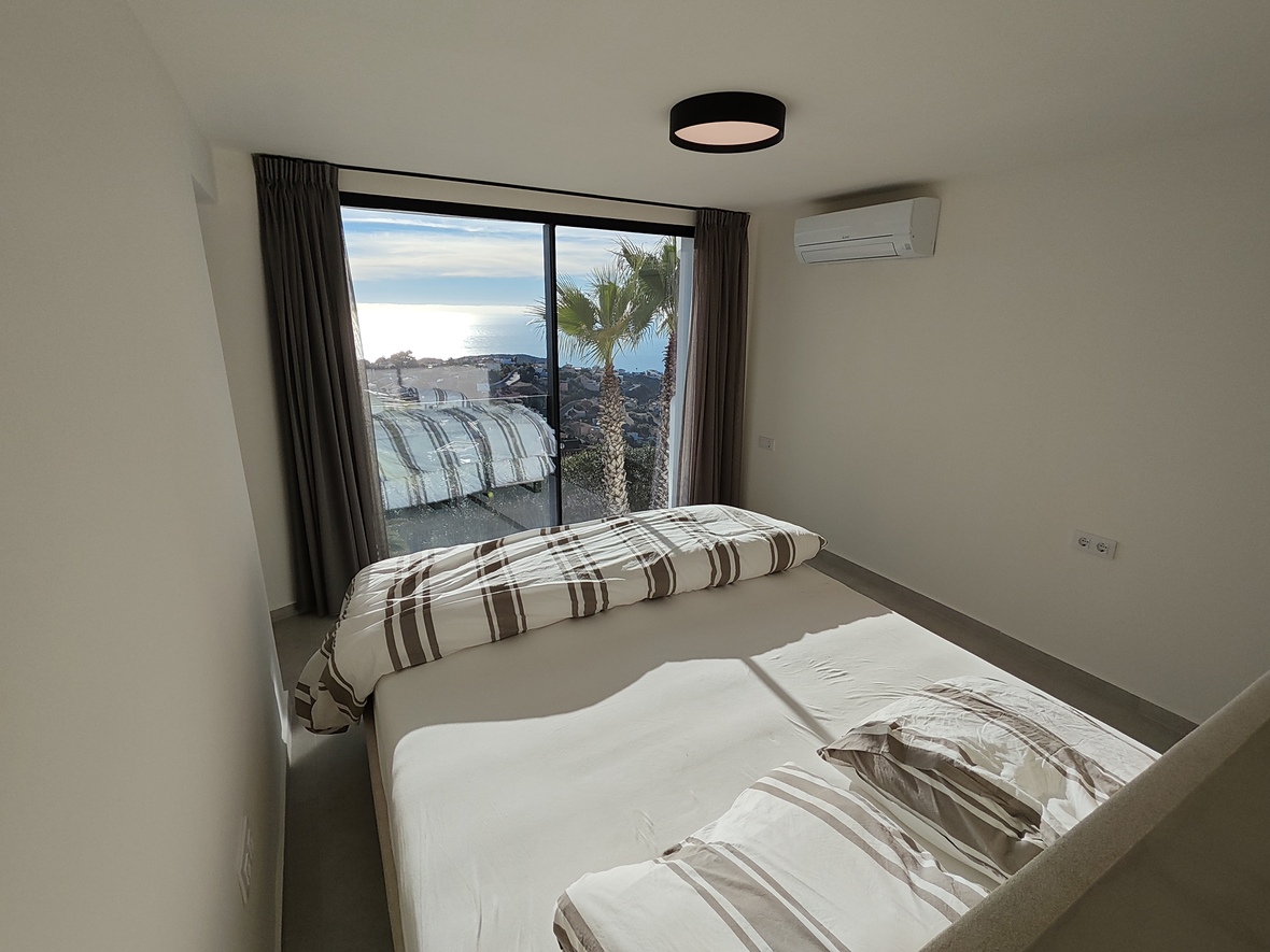 Chambre avec baies vitrées coulissantes offrant une vue sur la mer et les palmiers. Murs blancs minimalistes, literie à rayures et climatisation murale installée.
