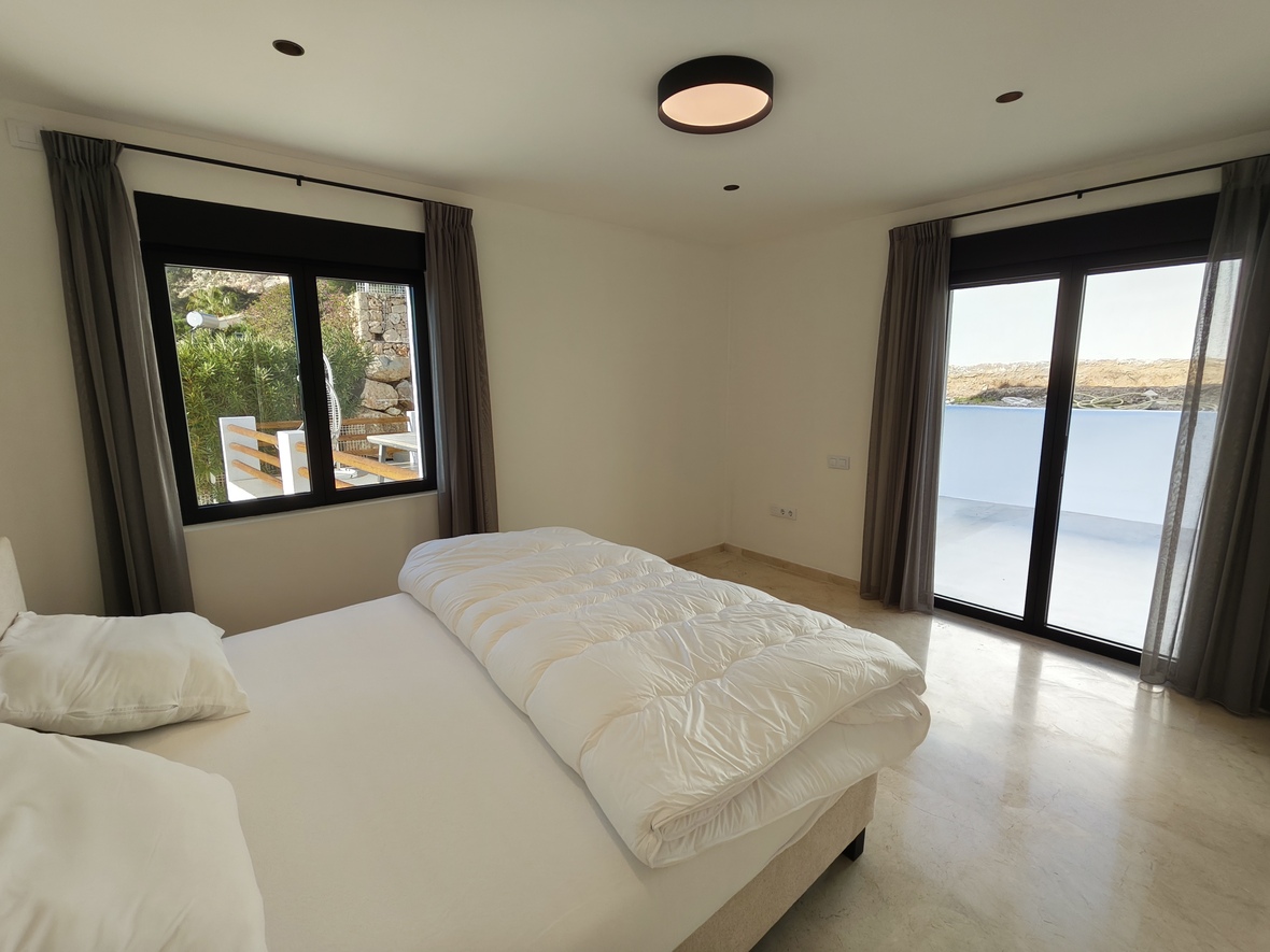 Chambre lumineuse avec sol en marbre, baies vitrées à cadre noir ouvrant sur une terrasse et rideaux gris. Éclairage minimaliste et spots encastrés au plafond.