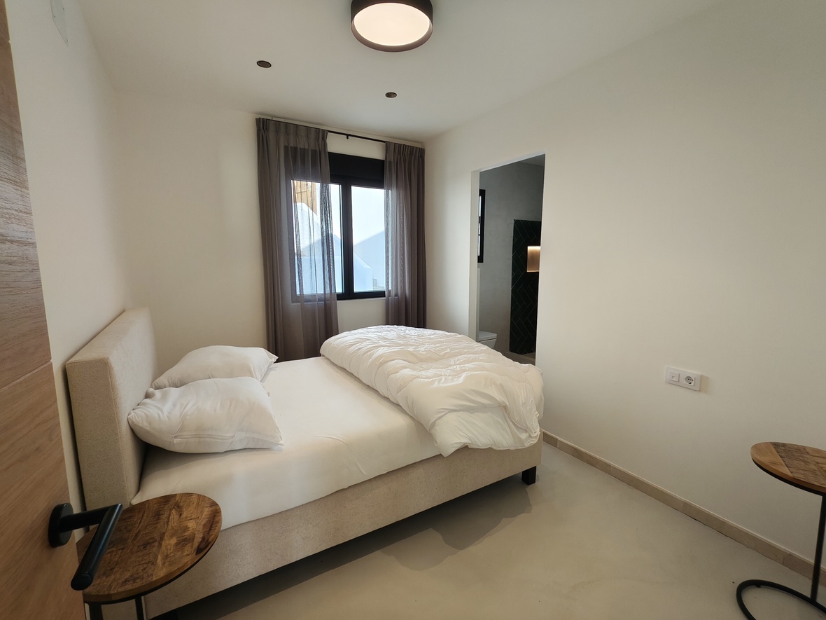 Chambre minimaliste avec lit rembourré beige, tables de chevet en bois et salle de bain attenante aux carreaux sombres. Sol en béton poli et fenêtres à cadre noir.