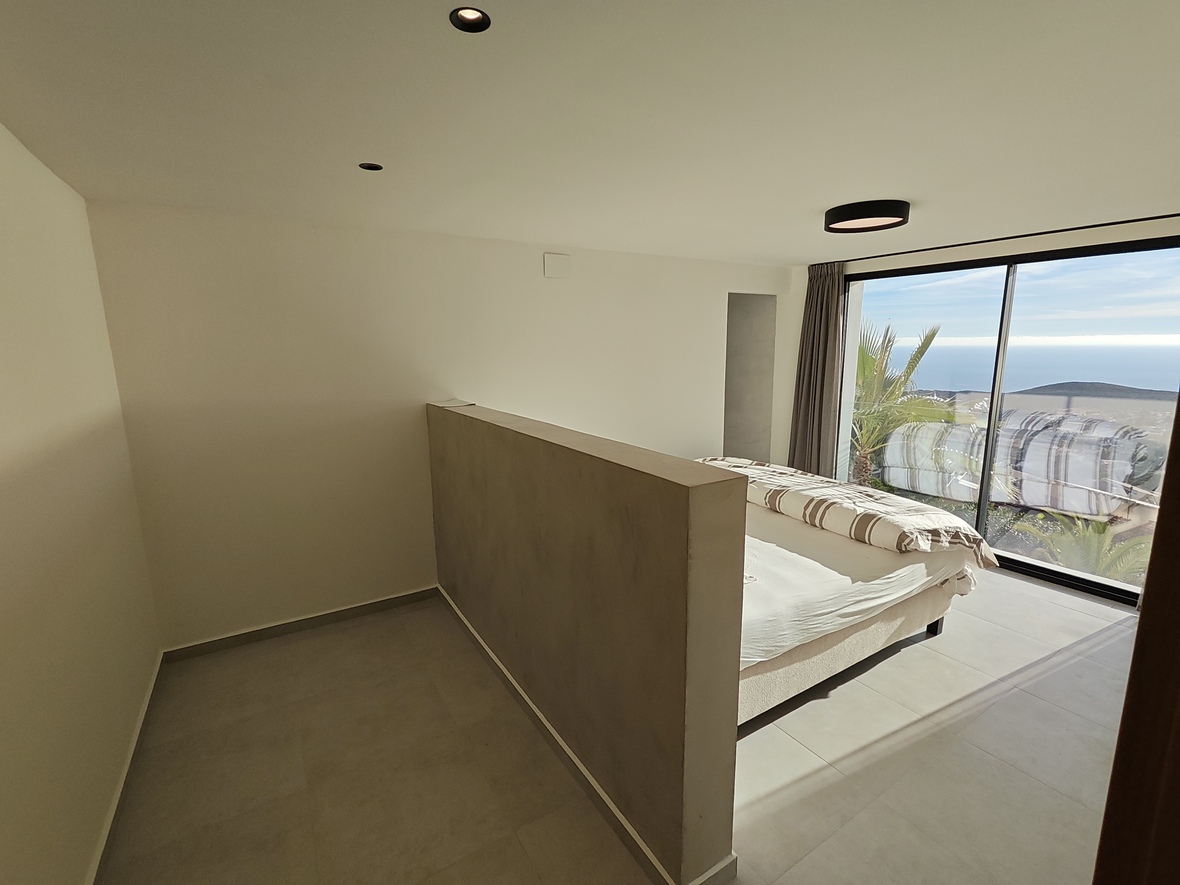 Chambre minimaliste avec muret de séparation, grandes baies vitrées offrant une vue sur l'océan et éclairage de plafond encastré.