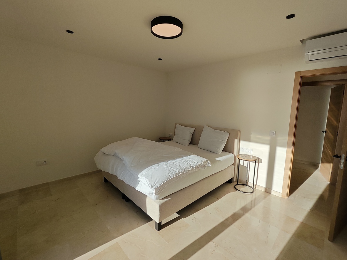 Chambre minimaliste avec sol en pierre polie, lit double rembourré et éclairage moderne comprenant un plafonnier circulaire et des spots encastrés.