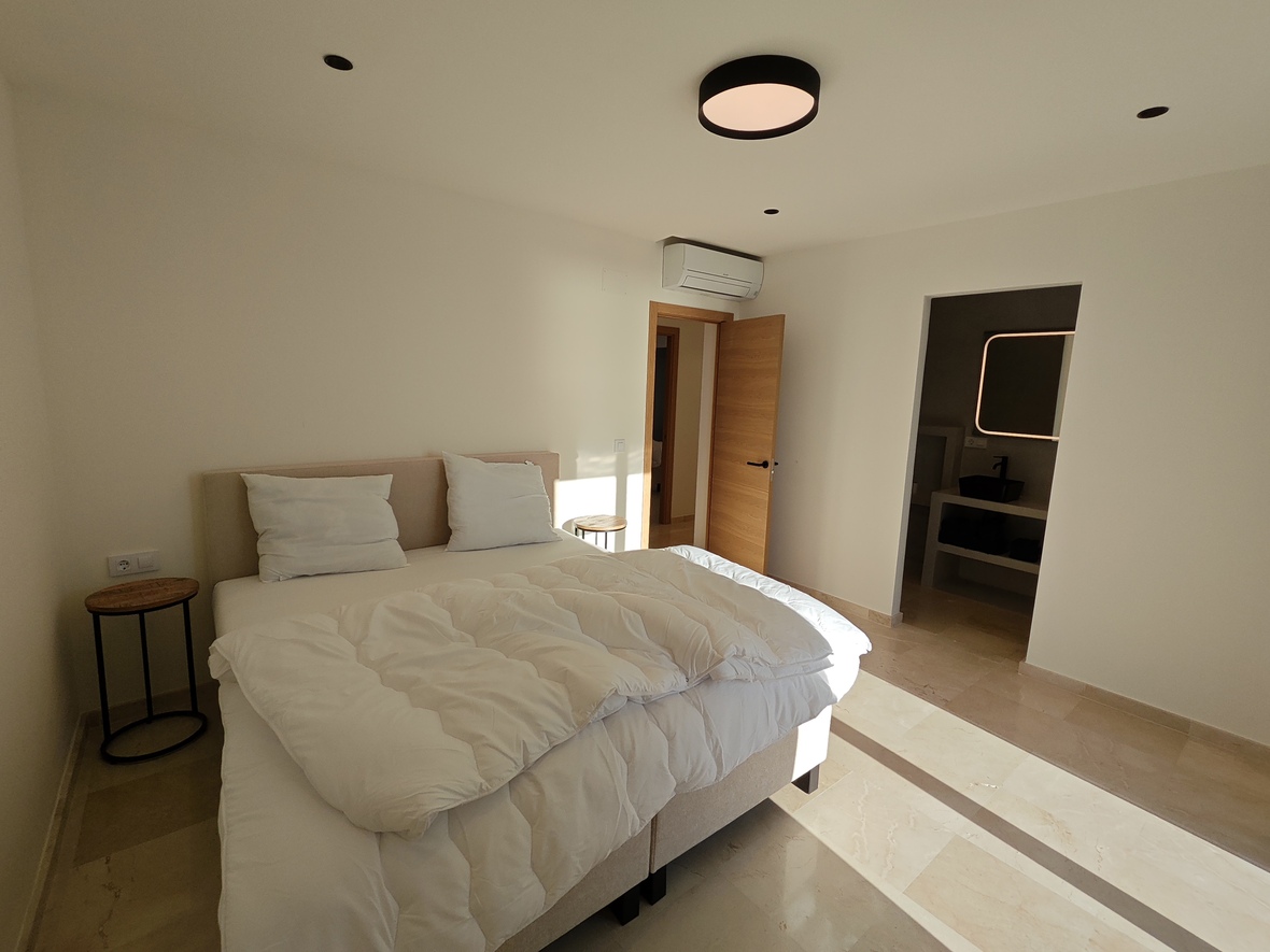 Chambre moderne avec sol en pierre, tête de lit rembourrée beige et tables de chevet minimalistes. Comprend la climatisation et une salle de bain attenante avec miroir rétroéclairé.