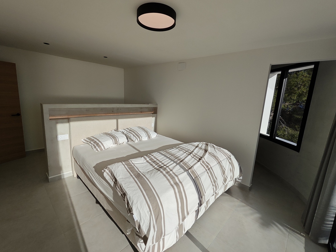 Chambre moderne avec tête de lit maçonnée intégrant un éclairage LED, carrelage grand format et fenêtre à cadre noir offrant une vue sur la végétation.