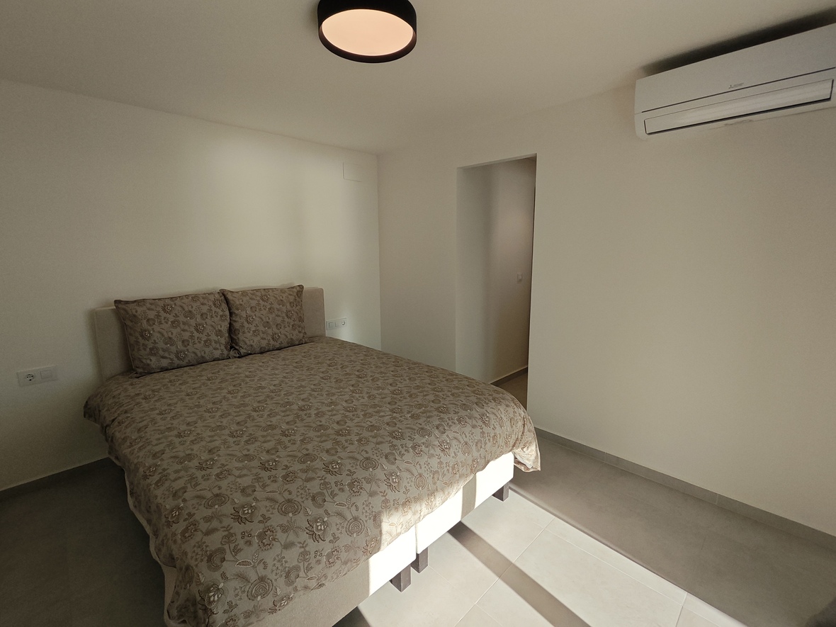 Chambre sobre avec lit double aux motifs floraux, système de climatisation réversible et plafonnier circulaire noir sur carrelage clair.