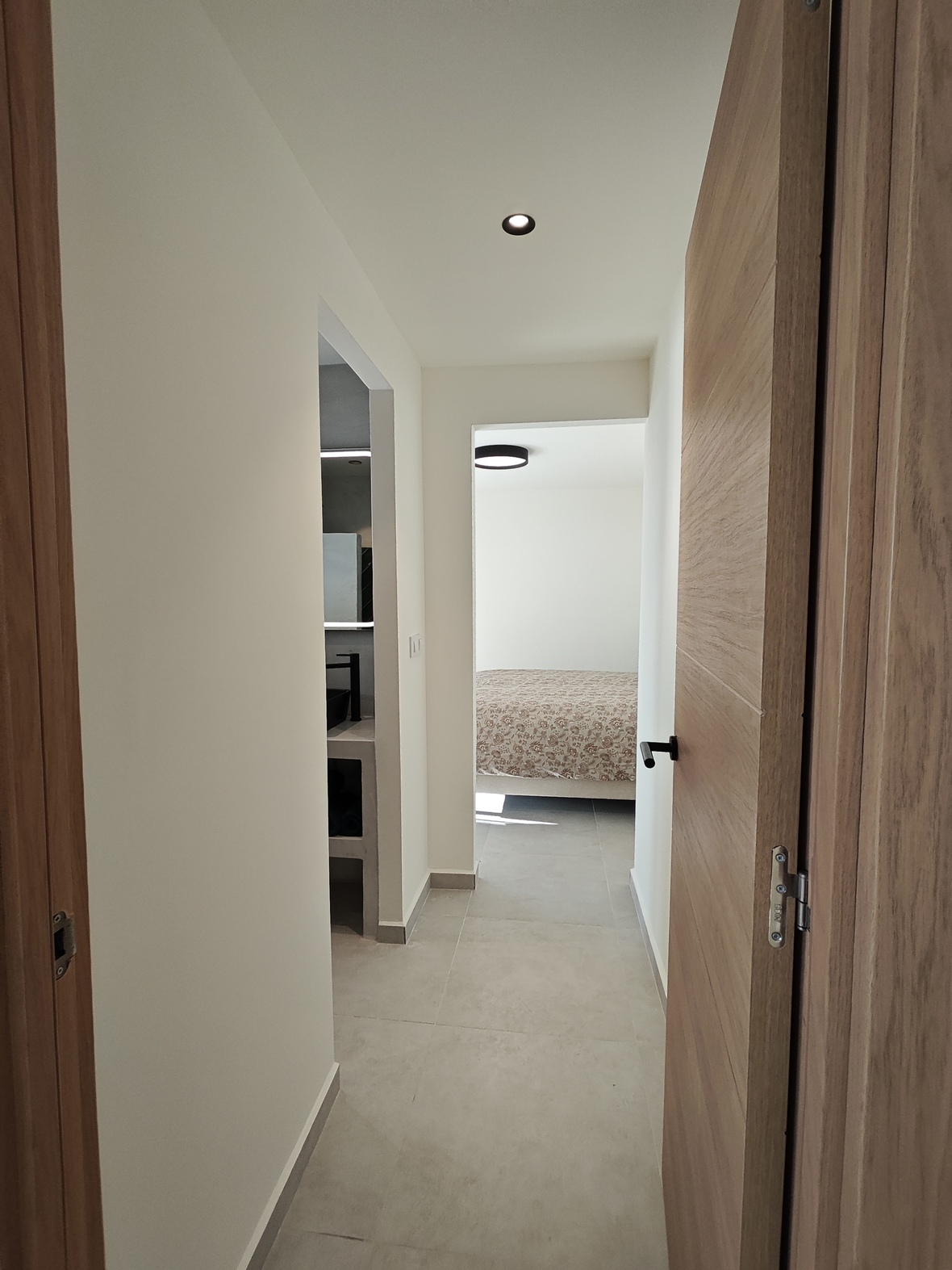 Couloir moderne avec portes en bois clair et sol en grès cérame gris, menant à une salle de bain minimaliste et une chambre lumineuse.
