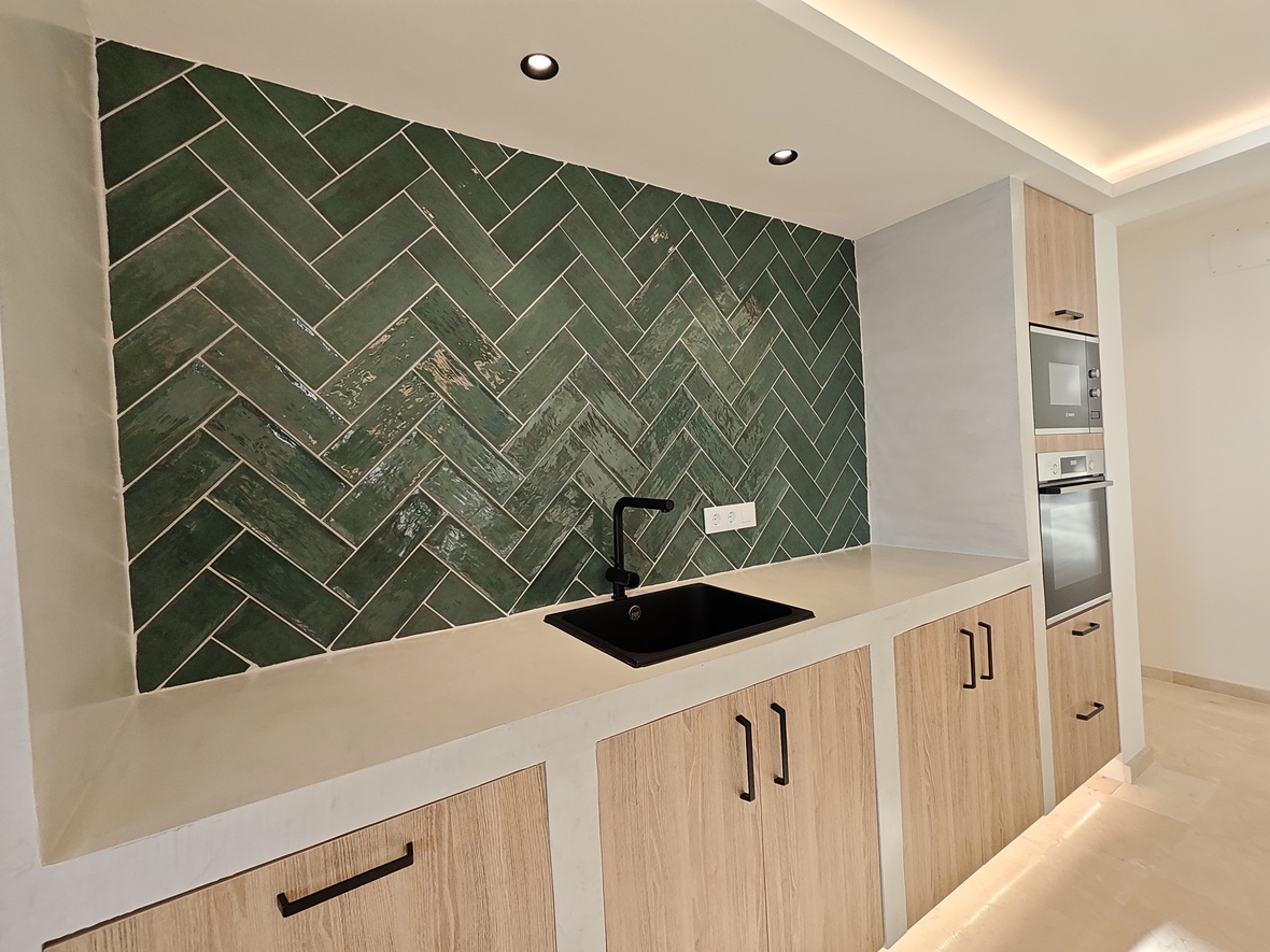 Cuisine moderne avec crédence en carreaux verts à chevrons, évier noir et placards en bois clair avec électroménager encastré et éclairage indirect.