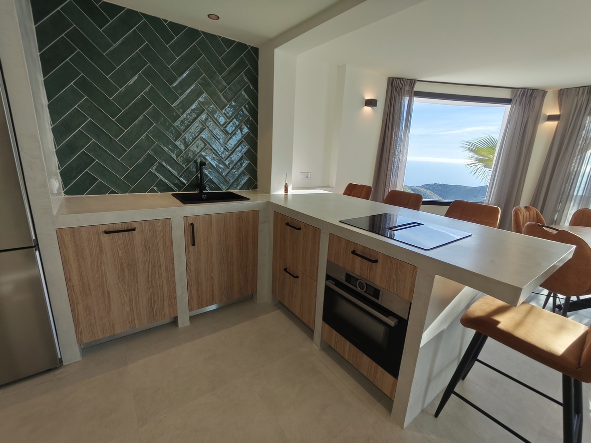 Cuisine moderne avec crédence en chevrons verts, meubles en bois et îlot central offrant une vue panoramique sur la mer et les montagnes.
