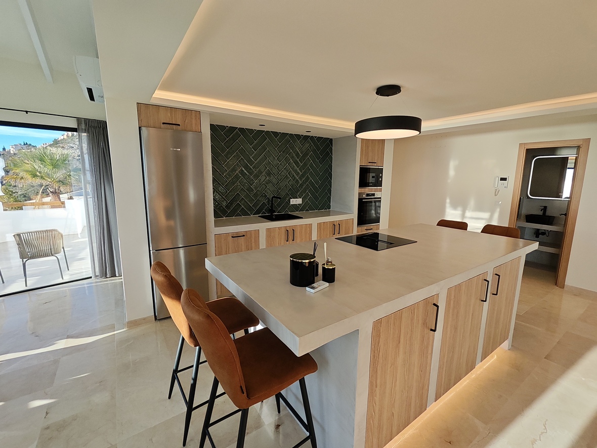 Cuisine moderne avec îlot central, plan de travail en pierre, tabourets en cuir et crédence en chevrons verts. Électroménagers intégrés et accès direct à la terrasse.
