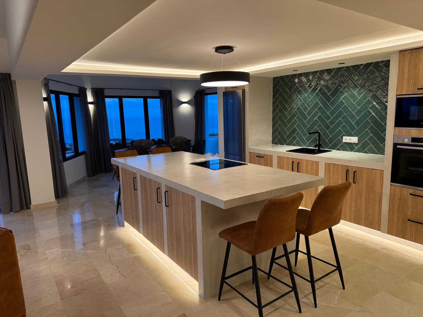 Cuisine moderne avec îlot en microciment, crédence en chevrons verts et vue panoramique sur la mer à travers de grandes baies vitrées au crépuscule.