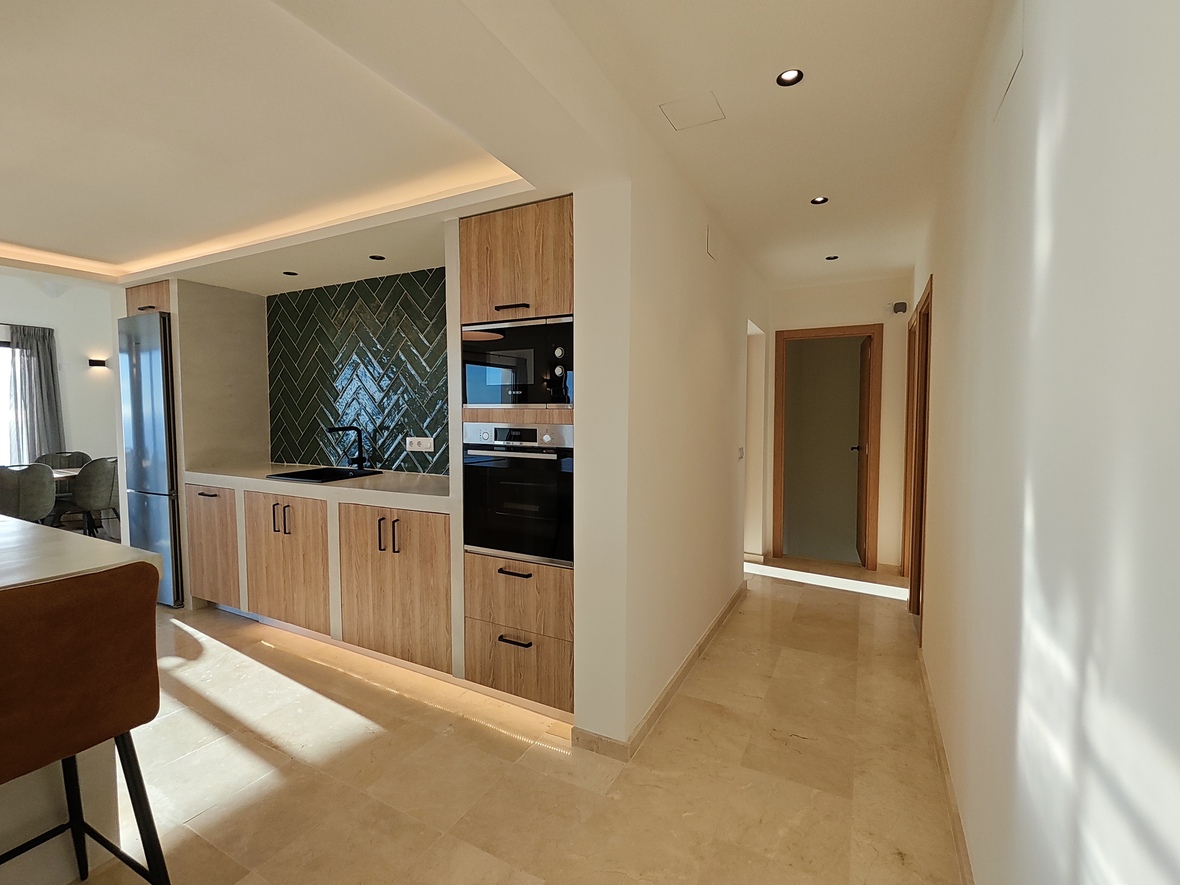 Cuisine moderne avec placards en bois, crédence en chevrons vert foncé et électroménager intégré, attenante à un couloir lumineux au sol en pierre naturelle.