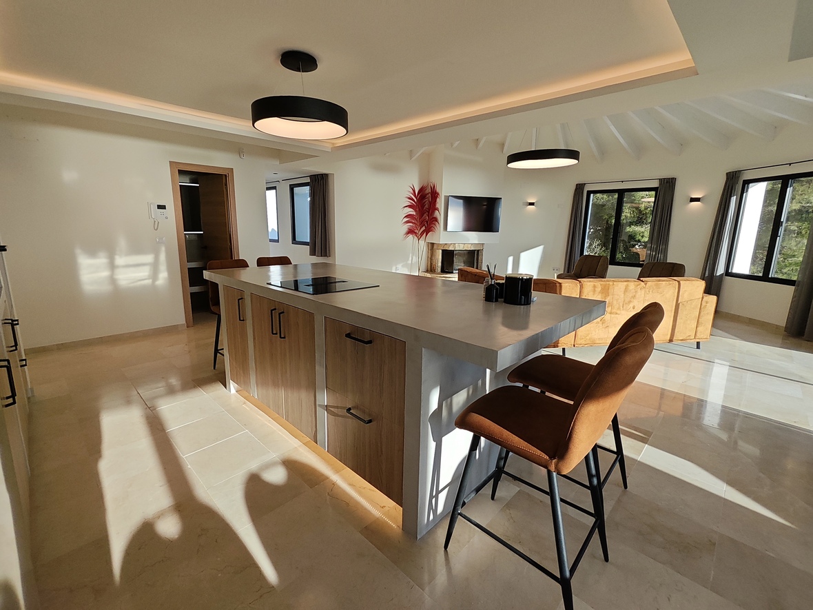 Cuisine ouverte avec îlot aspect béton, plaque à induction et tabourets en cuir, sous un plafond voûté à poutres blanches avec un éclairage circulaire moderne.