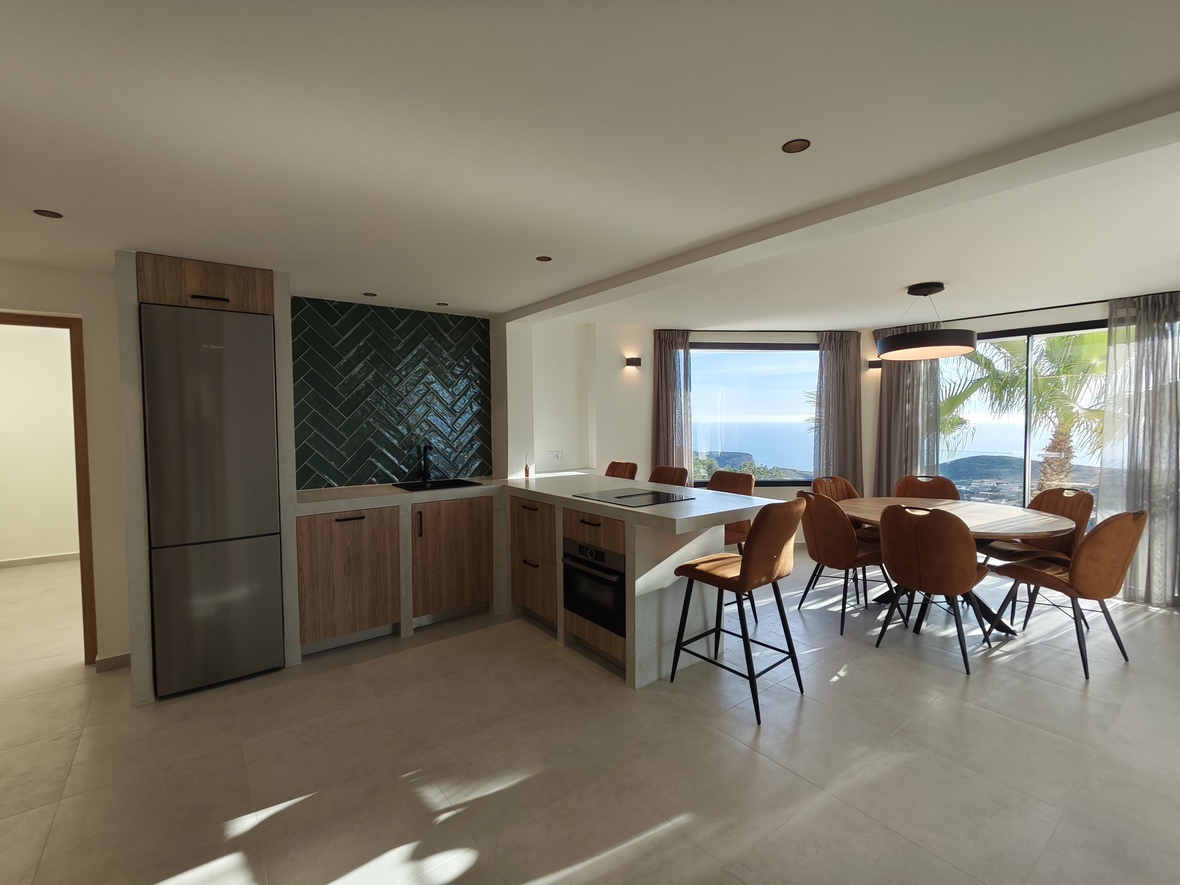Cuisine ouverte moderne avec crédence en chevrons verts, appareils intégrés et coin repas offrant une vue panoramique sur la mer et les palmiers.