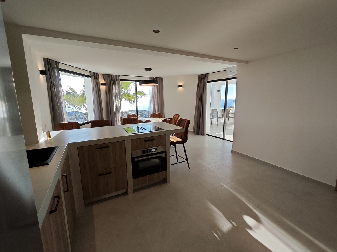 Cuisine ouverte moderne avec meubles en bois, îlot central et coin repas lumineux offrant une vue dégagée sur la mer et un accès à la terrasse extérieure.