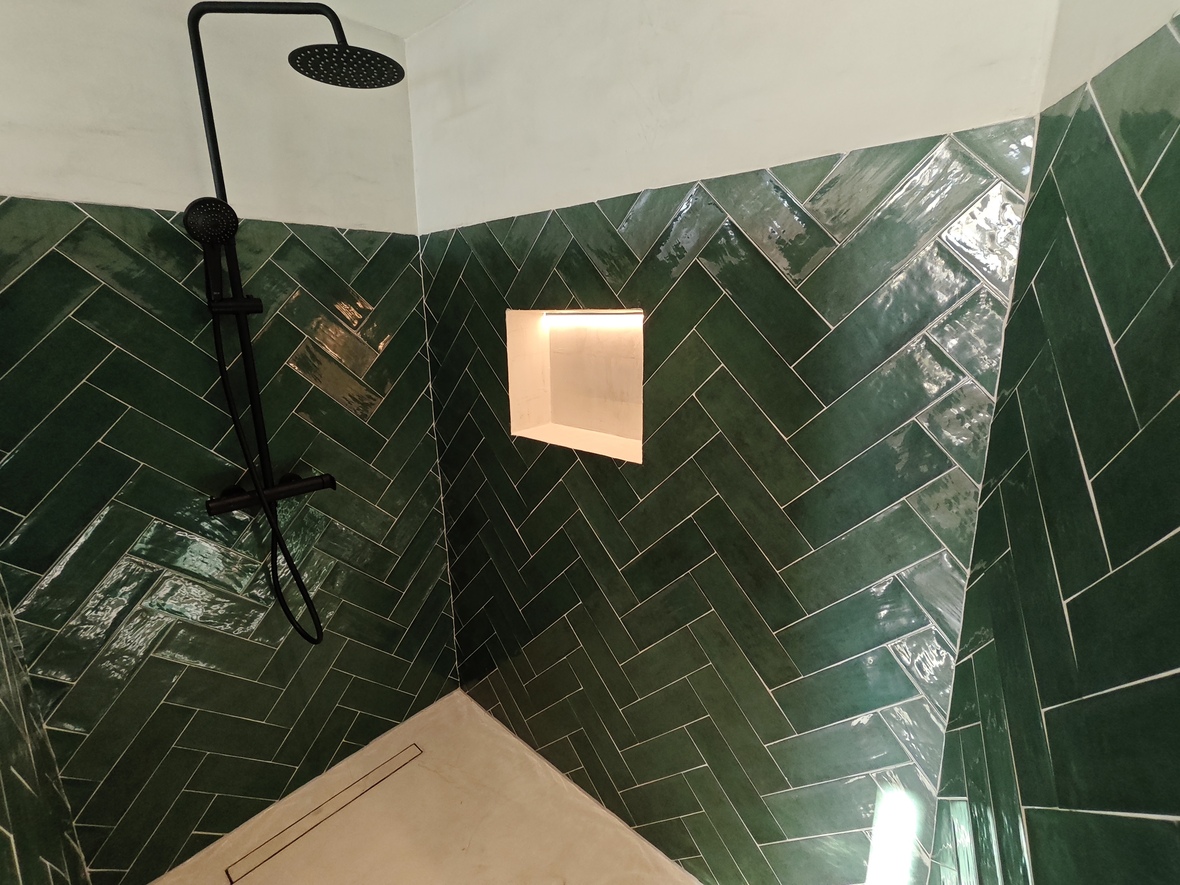 Douche avec carrelage vert émeraude en chevrons, robinetterie noire mate et niche murale éclairée par LED. Sol clair avec caniveau de douche linéaire.