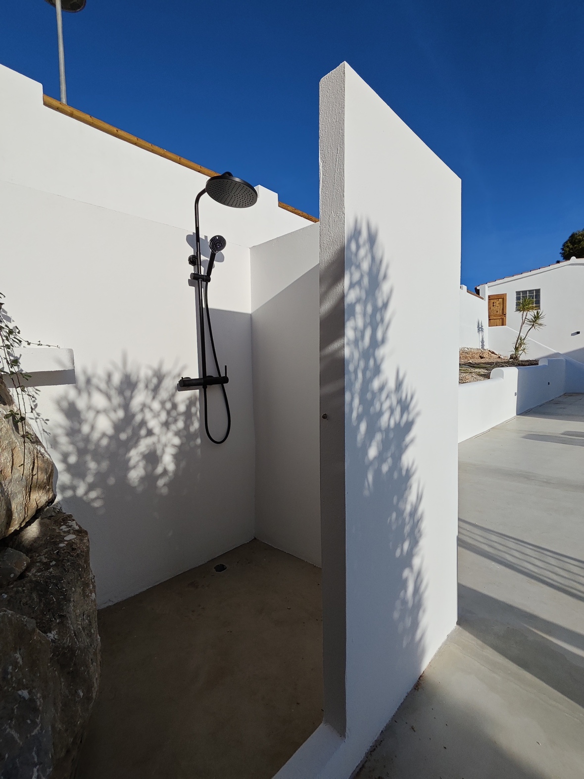 Douche extérieure minimaliste avec robinetterie noire mate et murs blancs, installée sur un sol en béton lissé sous un ciel azur.