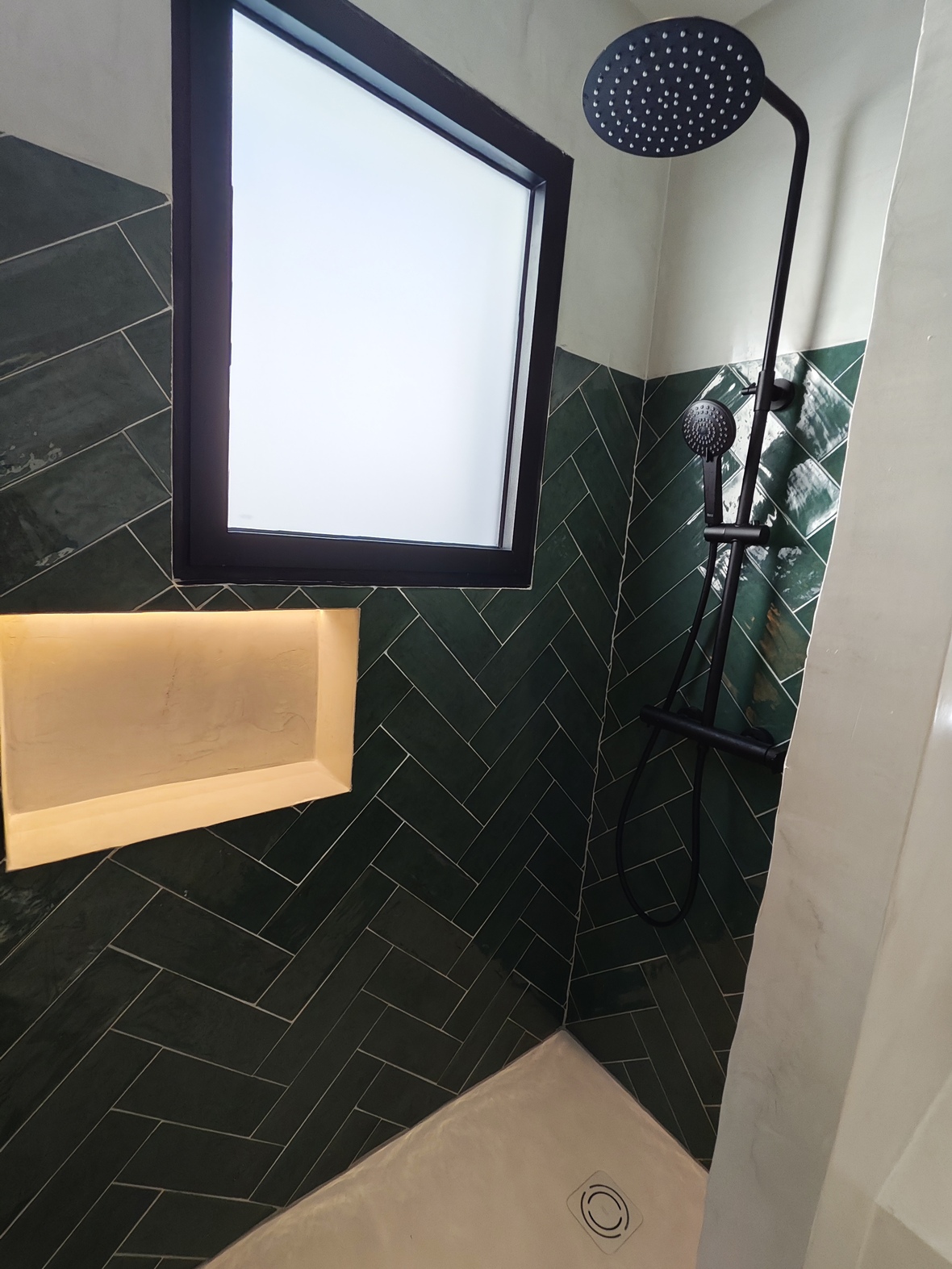 Douche moderne avec carrelage vert foncé en chevrons, robinetterie noire mate, niche éclairée et fenêtre en verre dépoli pour une lumière naturelle tamisée.