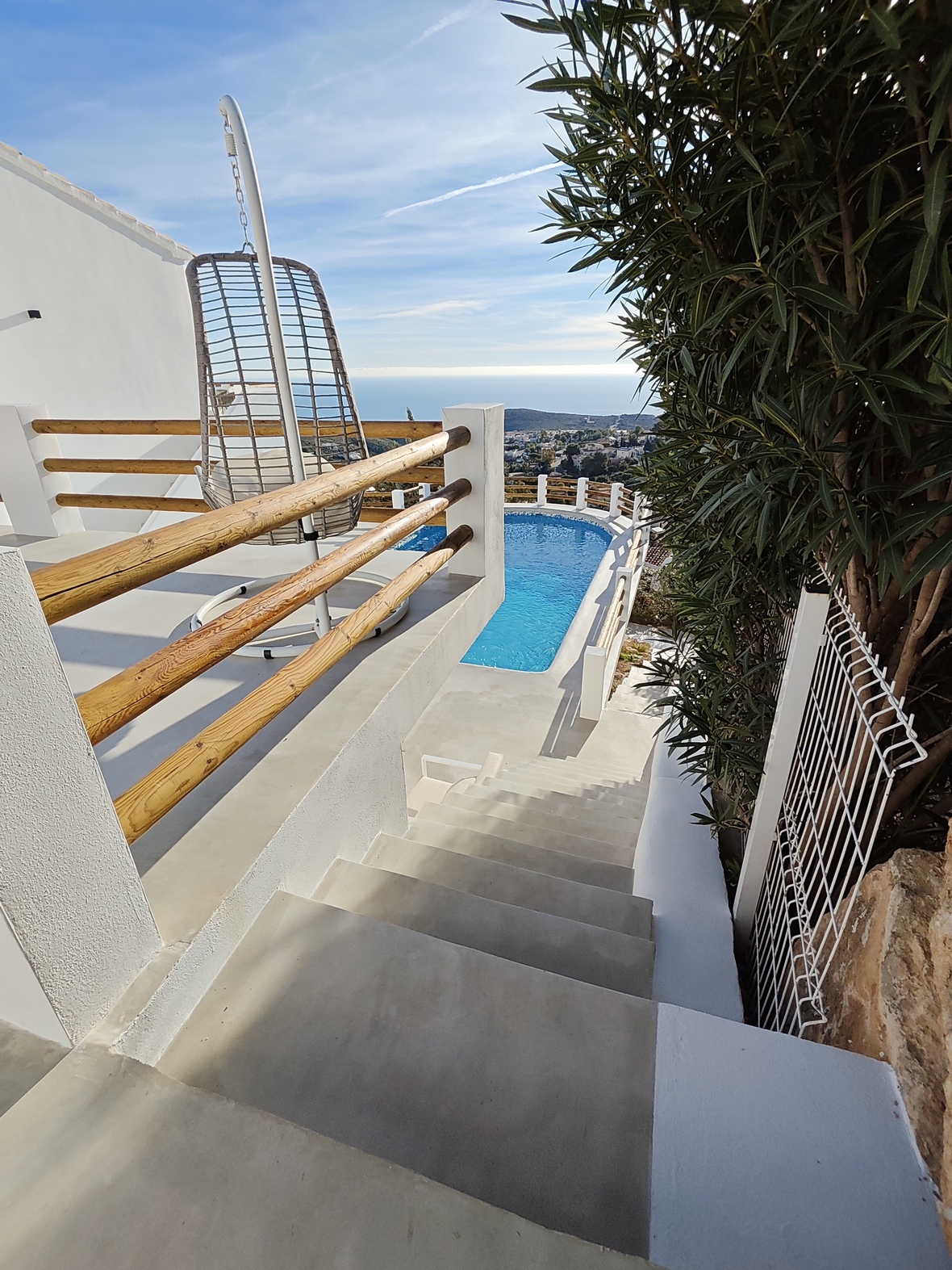 Escalier en béton menant à une terrasse avec fauteuil suspendu et balustrades en bois, offrant une vue sur la piscine et le littoral méditerranéen.