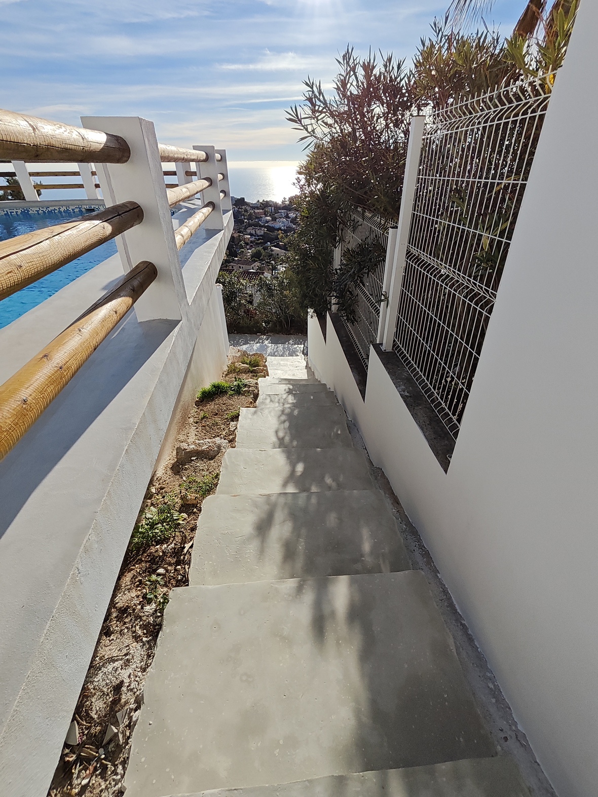 Escalier extérieur en béton avec garde-corps en bois et murs blancs, offrant une vue sur la piscine et le littoral méditerranéen ensoleillé.
