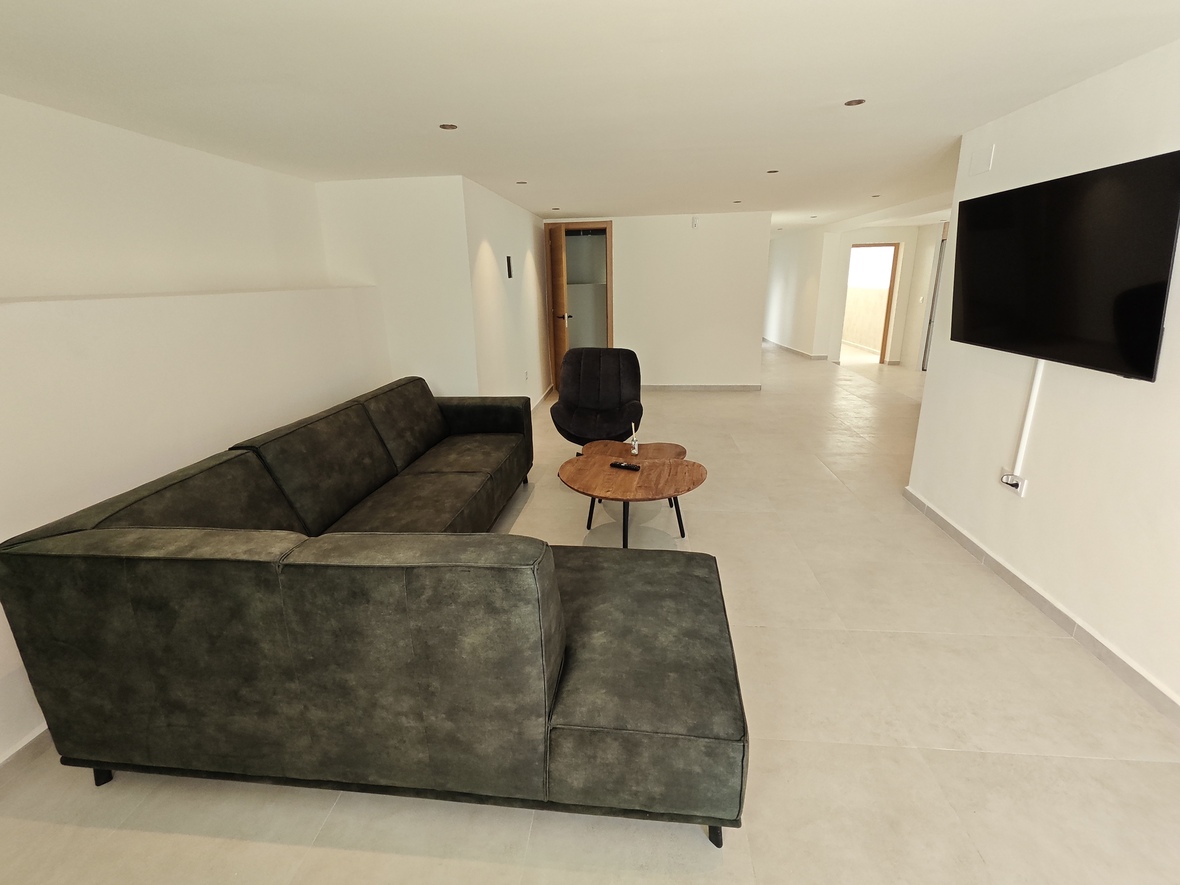Espace de vie moderne avec canapé d'angle vert foncé, table basse en bois naturel et téléviseur mural sur fond de murs blancs et sol carrelé clair.