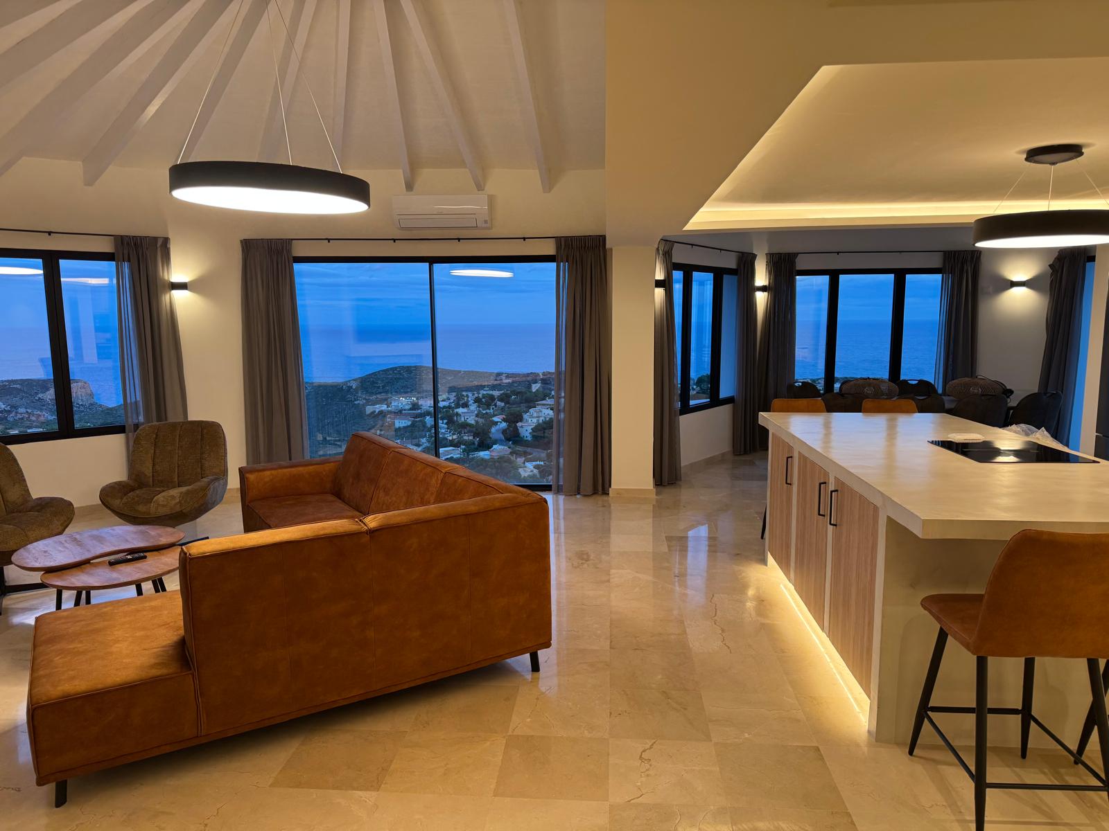 Espace de vie ouvert avec canapé en cuir cognac, îlot de cuisine avec éclairage LED et grandes baies vitrées offrant une vue panoramique sur la mer au crépuscule.