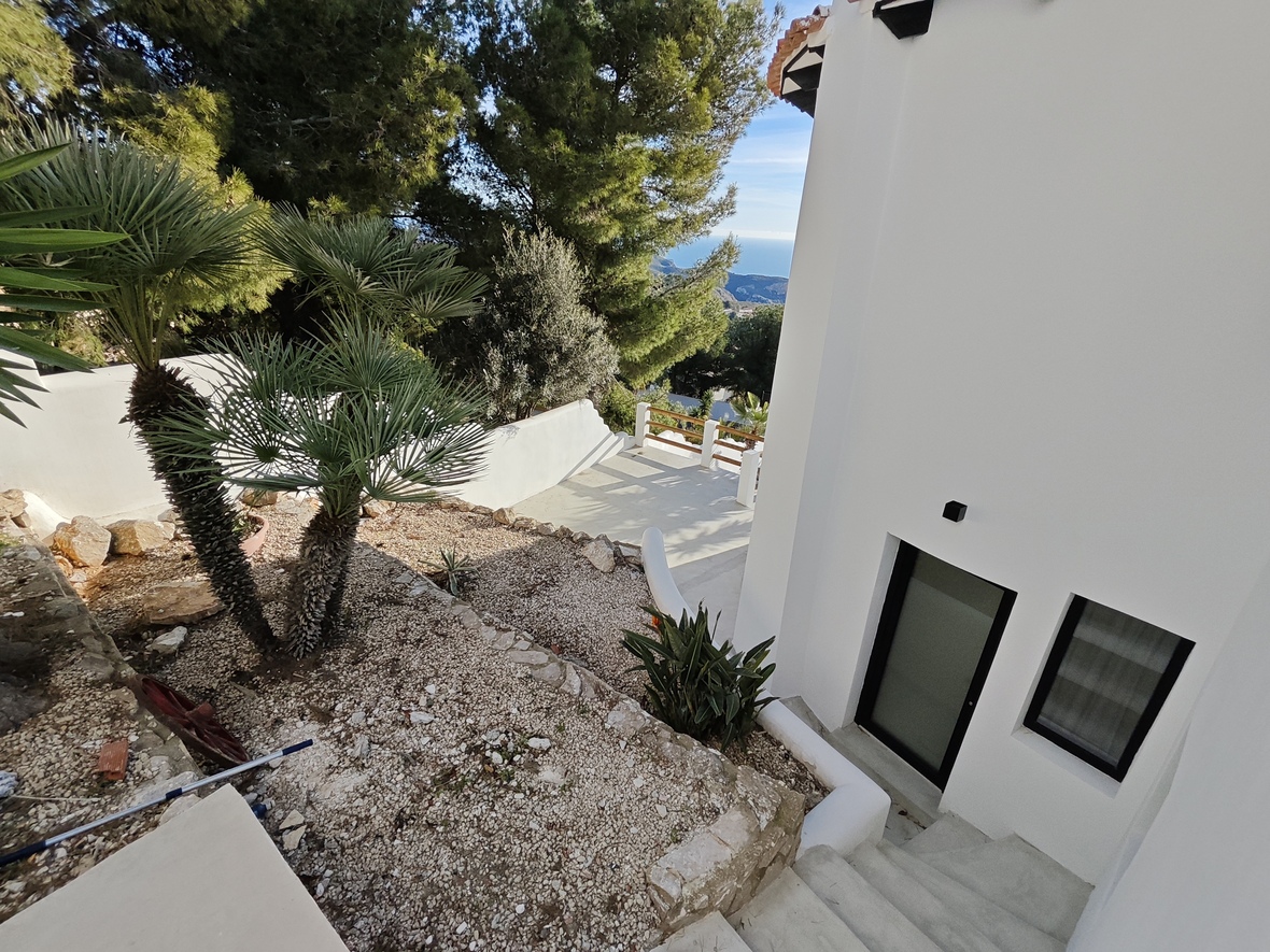 Extérieur de la villa avec jardin de gravier, palmiers et murs en pierre. Des escaliers mènent à une entrée moderne offrant une vue dégagée sur la mer.