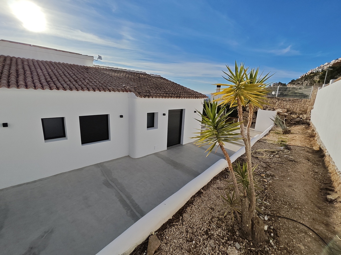 Extérieur d'une villa blanche minimaliste avec toit en tuiles, volets sombres et terrasse en béton bordée d'un yucca et d'un mur en pierre sèche.