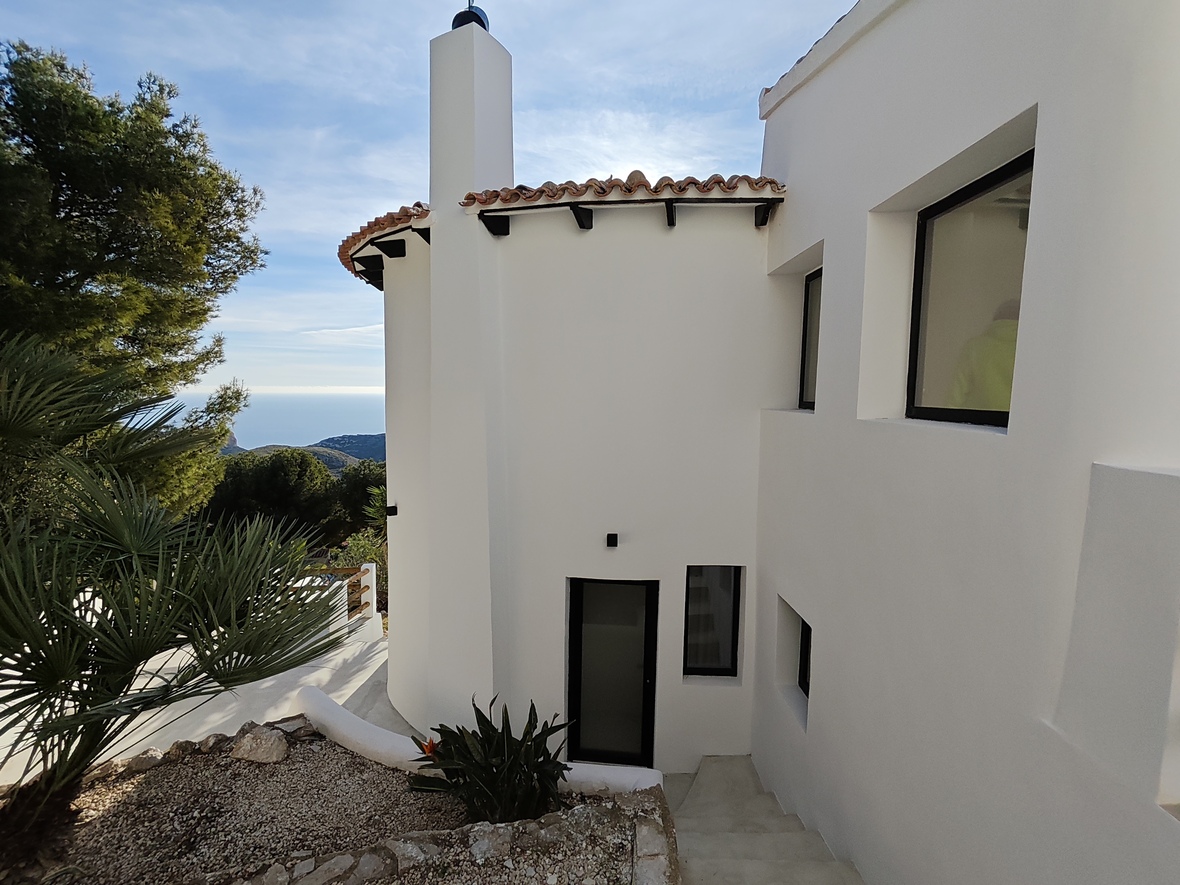 Extérieur d'une villa méditerranéenne aux murs blancs, toiture en terre cuite et escaliers menant à une entrée avec vue dégagée sur la mer et les montagnes.