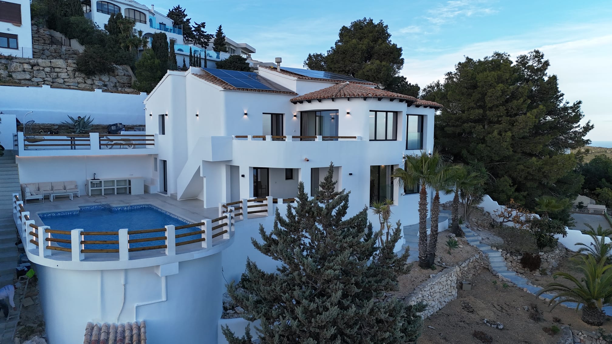 Façade d'une villa blanche à plusieurs niveaux avec piscine courbée, panneaux solaires et jardins en terrasses agrémentés de palmiers et de pins.