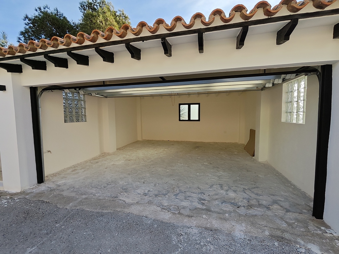 Garage spacieux avec sol en béton, briques de verre latérales pour la lumière naturelle et toiture traditionnelle en tuiles avec poutres apparentes sombres.