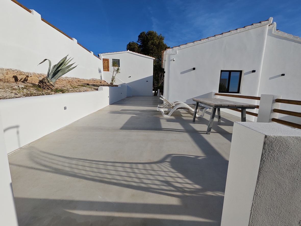 Grande terrasse extérieure avec sol en béton poli, bains de soleil blancs et table en bois, intégrée dans une architecture méditerranéenne aux murs blancs.