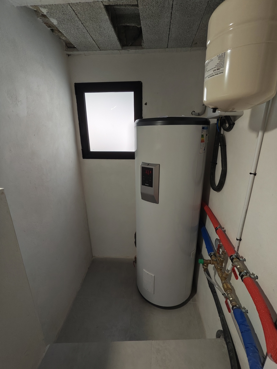 Local technique équipé d'un chauffe-eau thermodynamique Haier, d'un vase d'expansion et de tuyauteries isolées. Fenêtre dépolie et sol en grès gris.