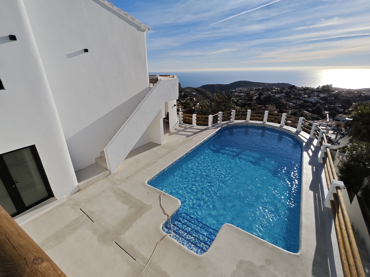 Piscine aux mosaïques bleues et terrasse en béton blanc avec garde-corps en bois, offrant une vue panoramique sur la mer Méditerranée et la côte.