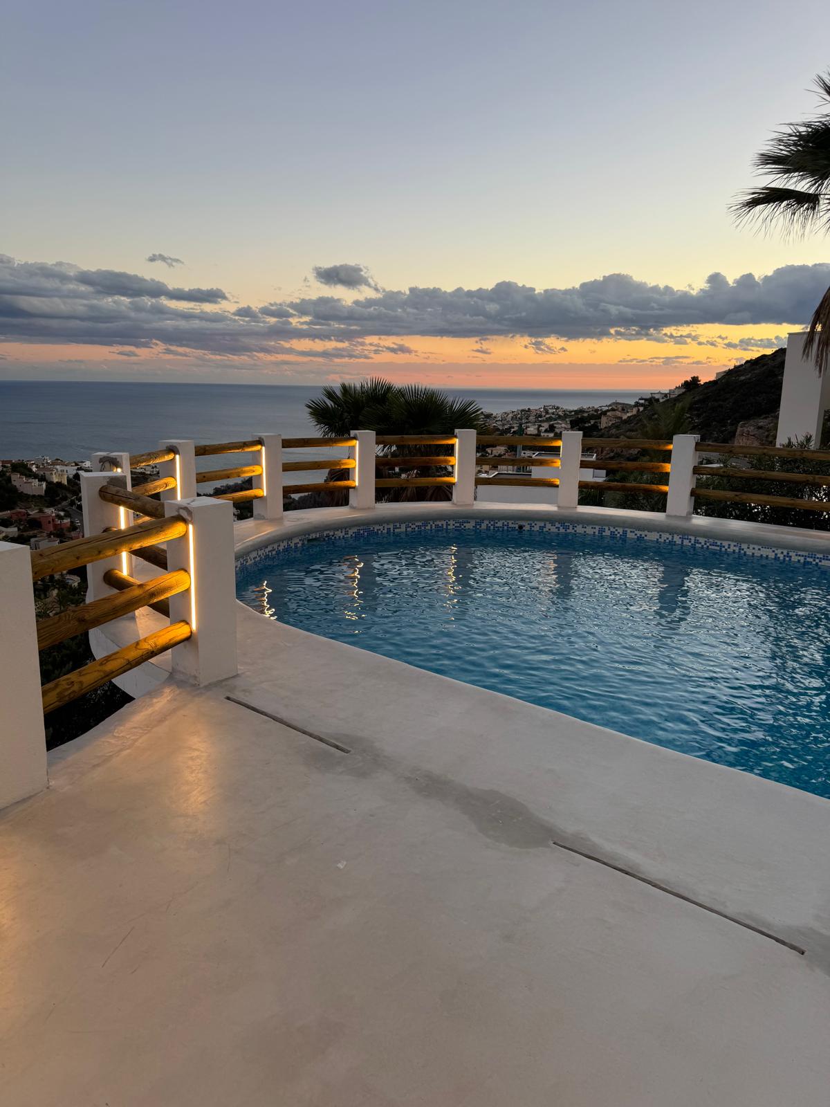 Piscine circulaire avec mosaïque bleue et éclairage LED intégré dans la balustrade en bois, offrant une vue panoramique sur la côte au crépuscule.