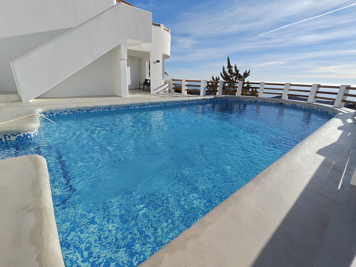 Piscine extérieure avec mosaïque bleue, terrasse en béton blanc et garde-corps en bois offrant une vue dégagée sur la mer.