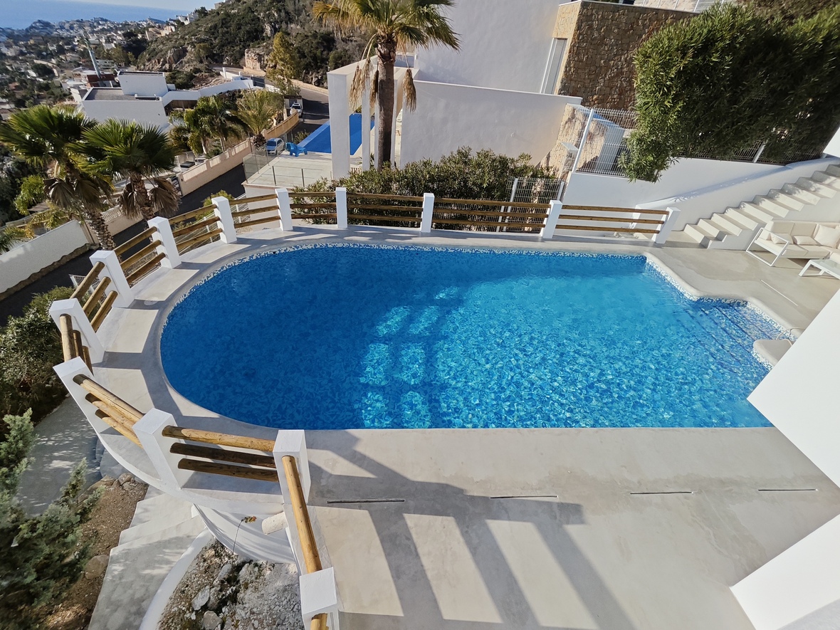 Piscine extérieure incurvée avec mosaïque bleue, entourée d'une terrasse en pierre et d'un garde-corps en bois offrant une vue panoramique sur la côte.
