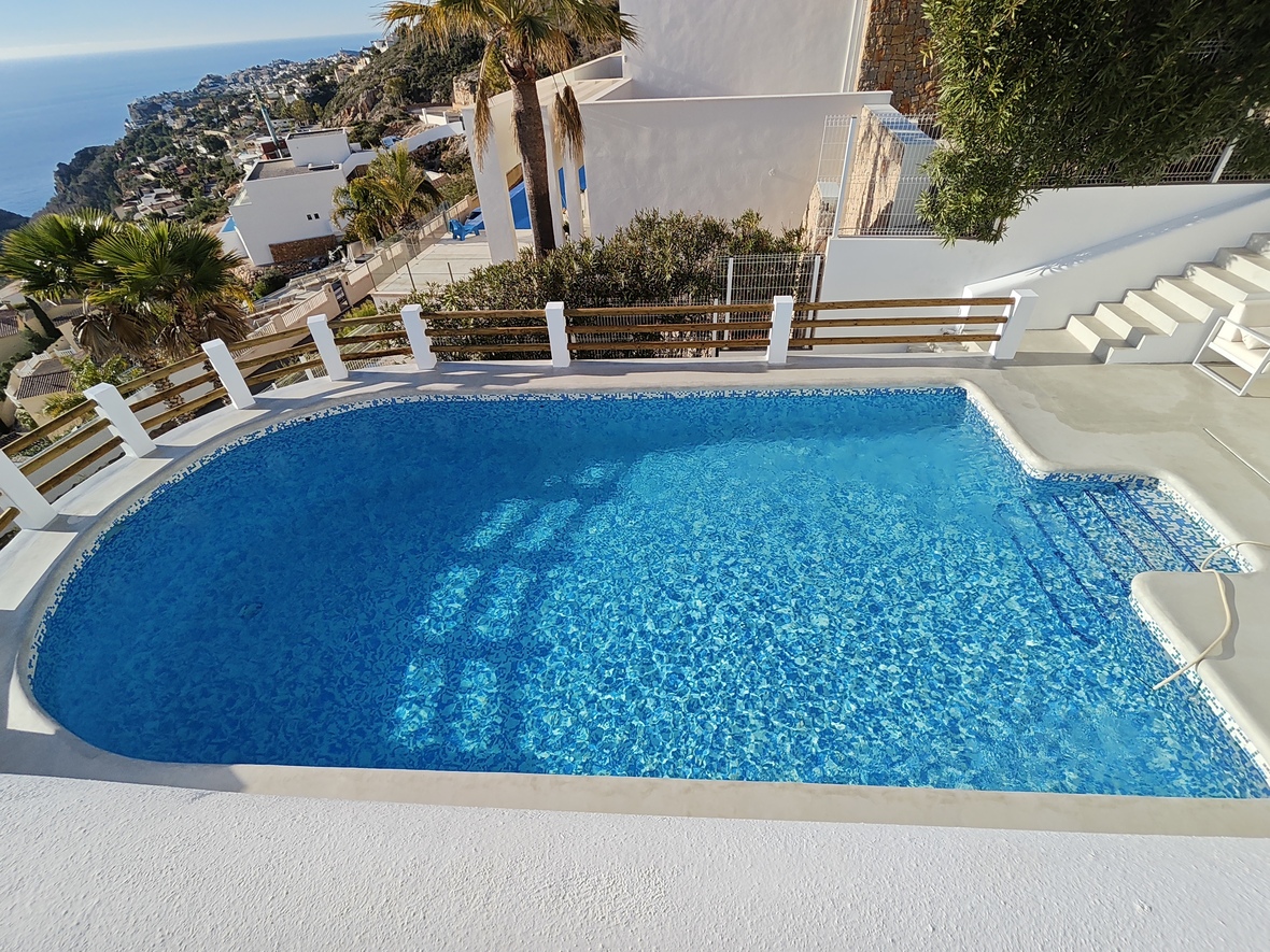 Piscine privée en mosaïque bleue avec escaliers intégrés, entourée d'une terrasse blanche et d'un garde-corps en bois offrant une vue dégagée sur la côte.