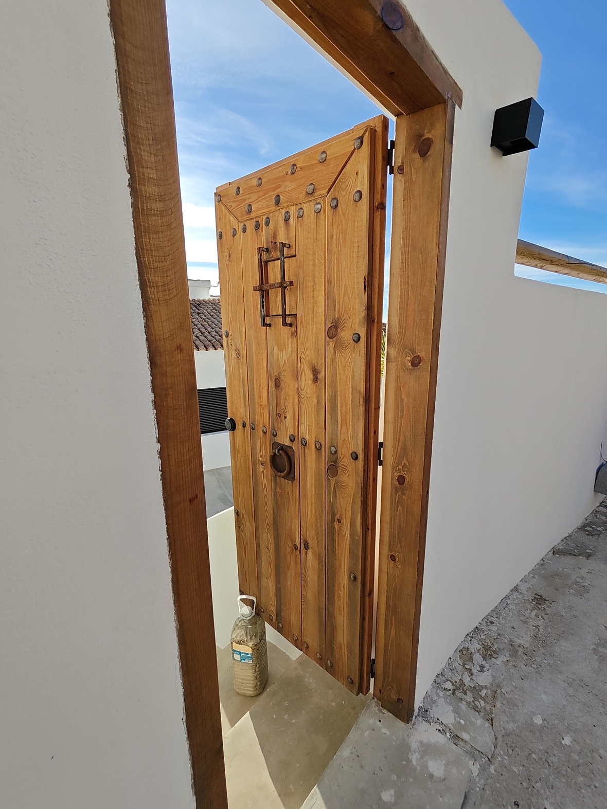 Porte traditionnelle en bois massif avec clous en fer et judas grillagé, encadrée dans un mur blanc épuré sous un ciel dégagé.