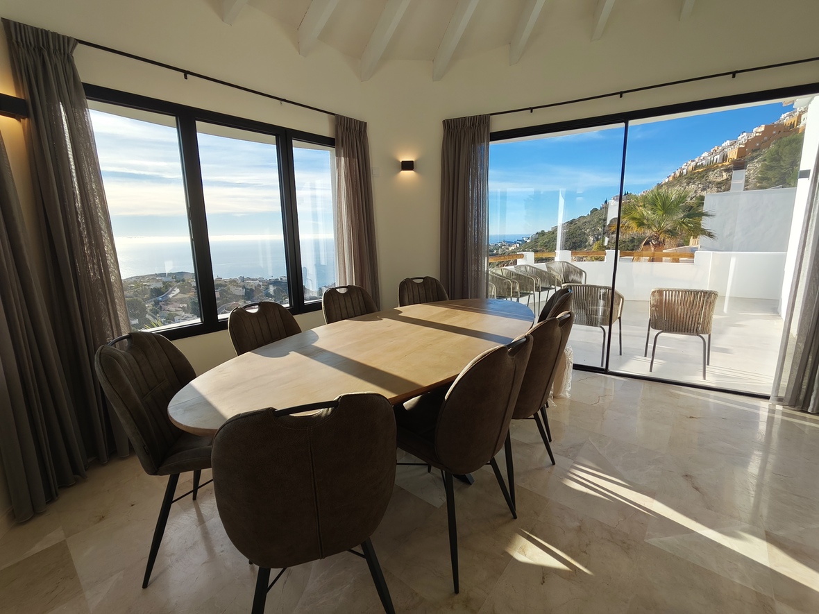 Salle à manger avec table ovale en bois et sol en marbre, offrant une vue panoramique sur la mer via de larges baies vitrées et un accès à la terrasse.