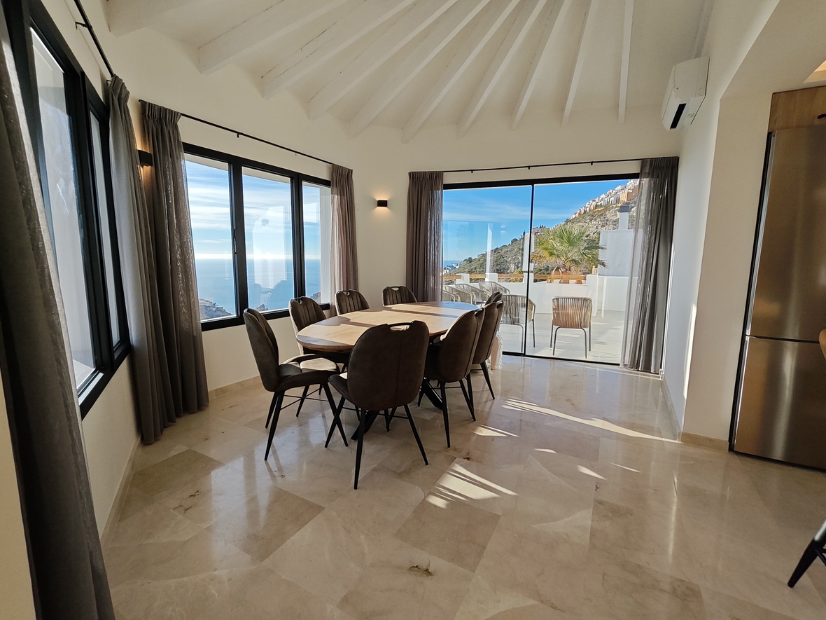 Salle à manger lumineuse avec table ovale en bois, sol en marbre et plafond voûté. Les baies vitrées offrent une vue sur la mer et un accès à la terrasse.