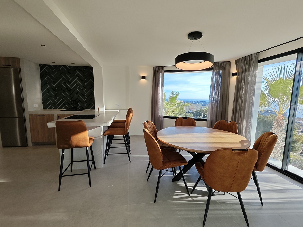 Salle à manger ouverte avec table ronde en bois, chaises cognac et cuisine moderne avec crédence en chevrons offrant une vue dégagée sur la mer.