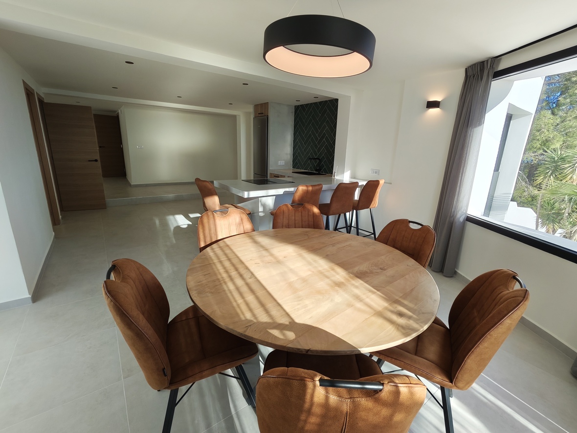 Salle à manger ouverte avec table ronde en bois, chaises couleur cognac et suspension design, attenante à une cuisine moderne avec îlot blanc et crédence verte.