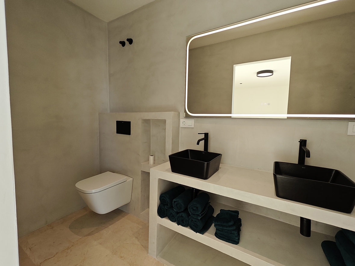 Salle de bain au design minimaliste avec murs en microciment gris, double vasque noire et un grand miroir à éclairage LED périphérique.