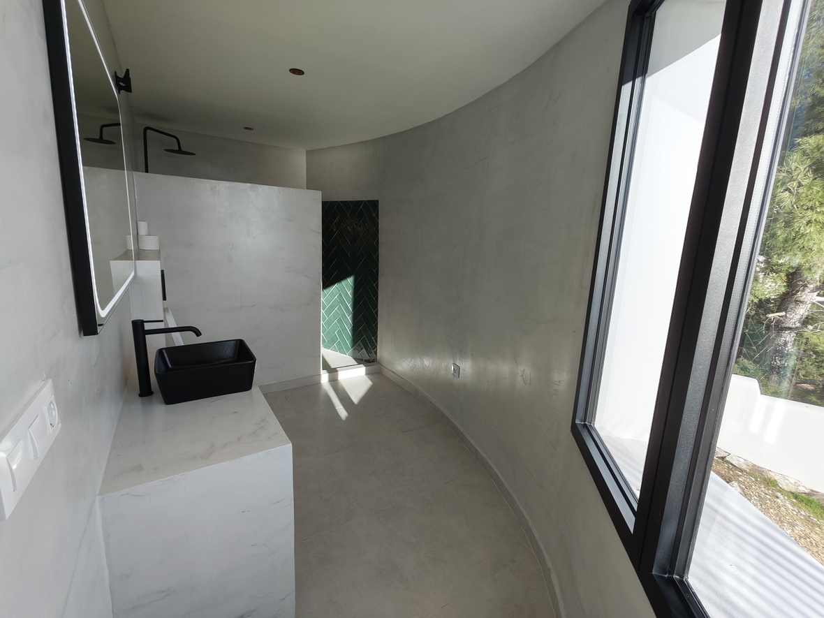 Salle de bain minimaliste aux murs incurvés en microciment, avec vasque noire et douche à l'italienne ornée de carreaux verts en chevrons.