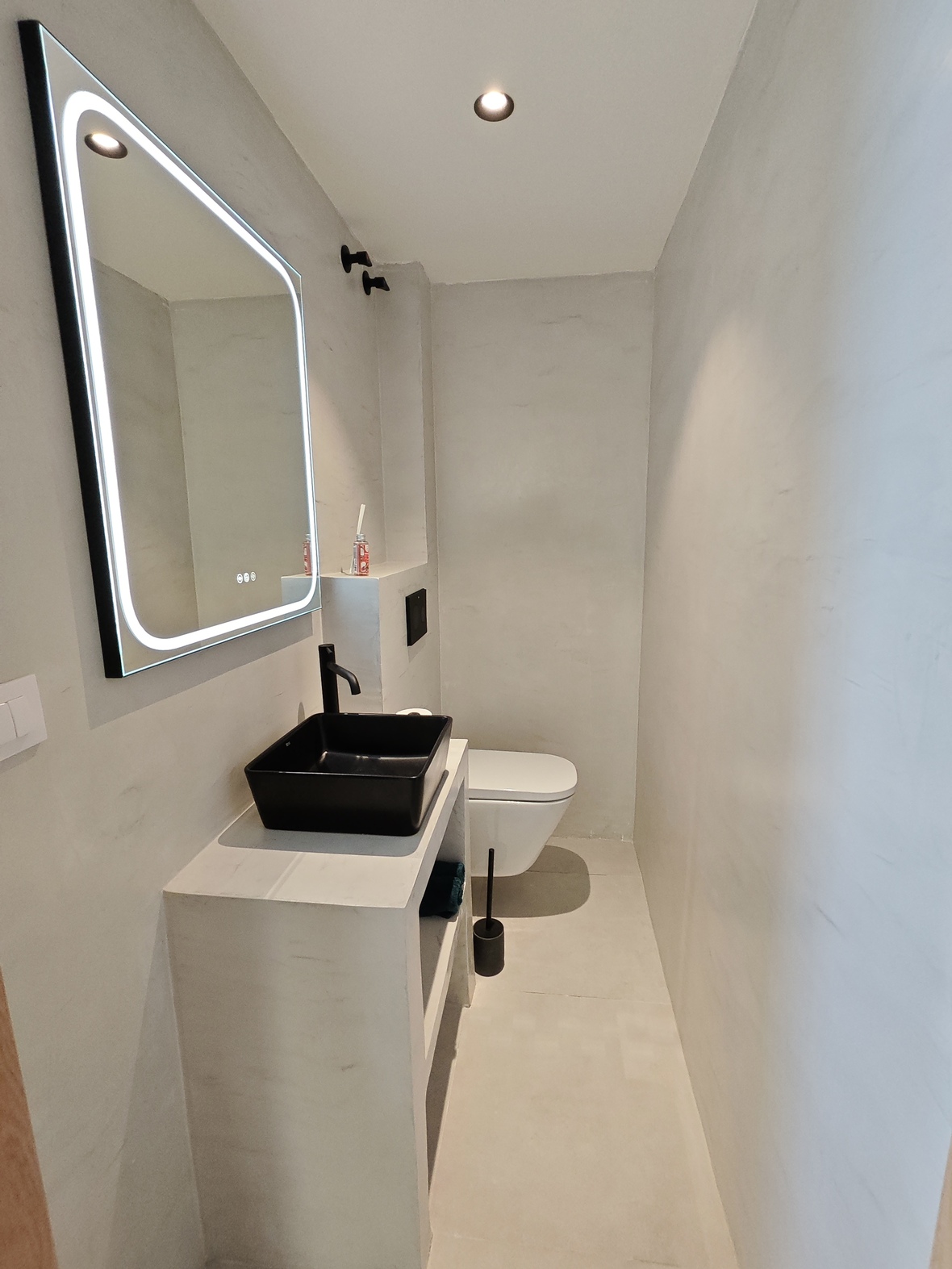 Salle de bain minimaliste avec vasque à poser noire, robinetterie mate, miroir rétroéclairé par LED et murs en microciment gris clair.