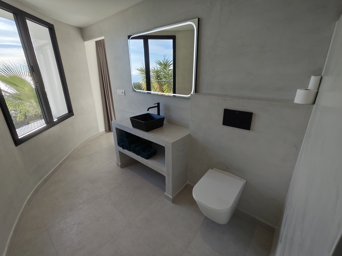 Salle de bain moderne avec murs en microciment, vasque noire et miroir rétroéclairé. Les grandes fenêtres apportent une lumière naturelle et des vues sur les palmiers.
