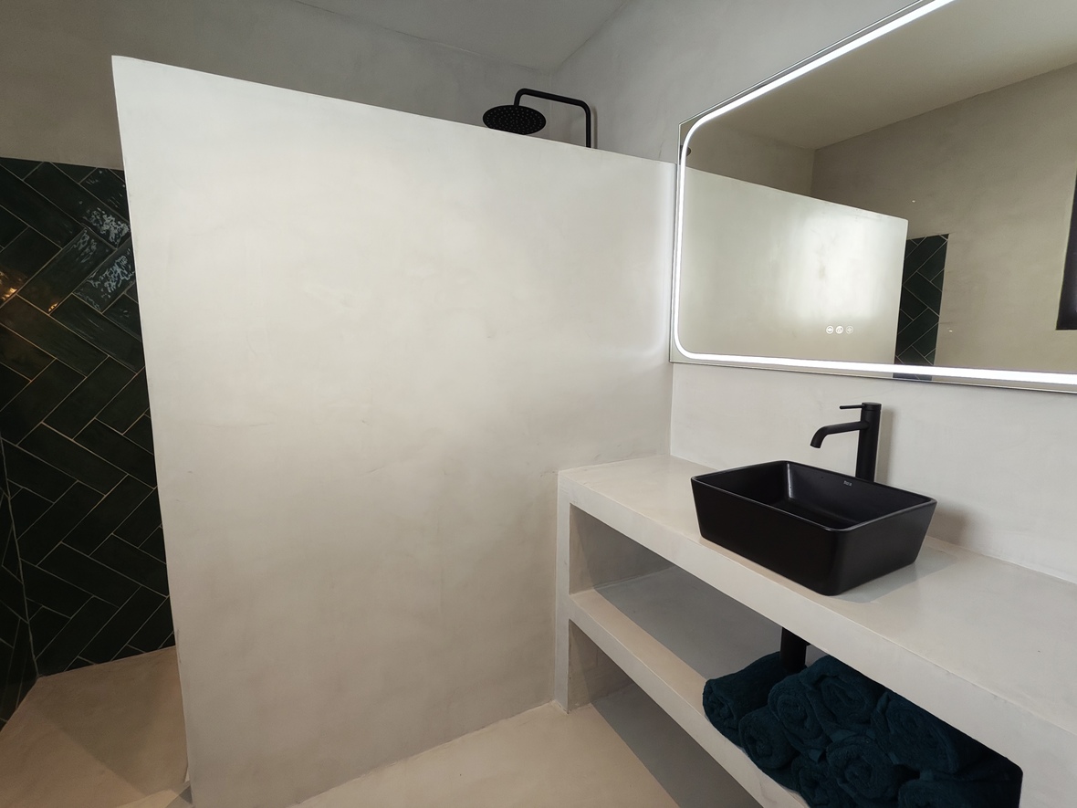 Salle de bain moderne en microciment avec vasque noire et miroir rétroéclairé. La douche à l'italienne présente des carreaux en chevrons vert foncé et une robinetterie noire mate.