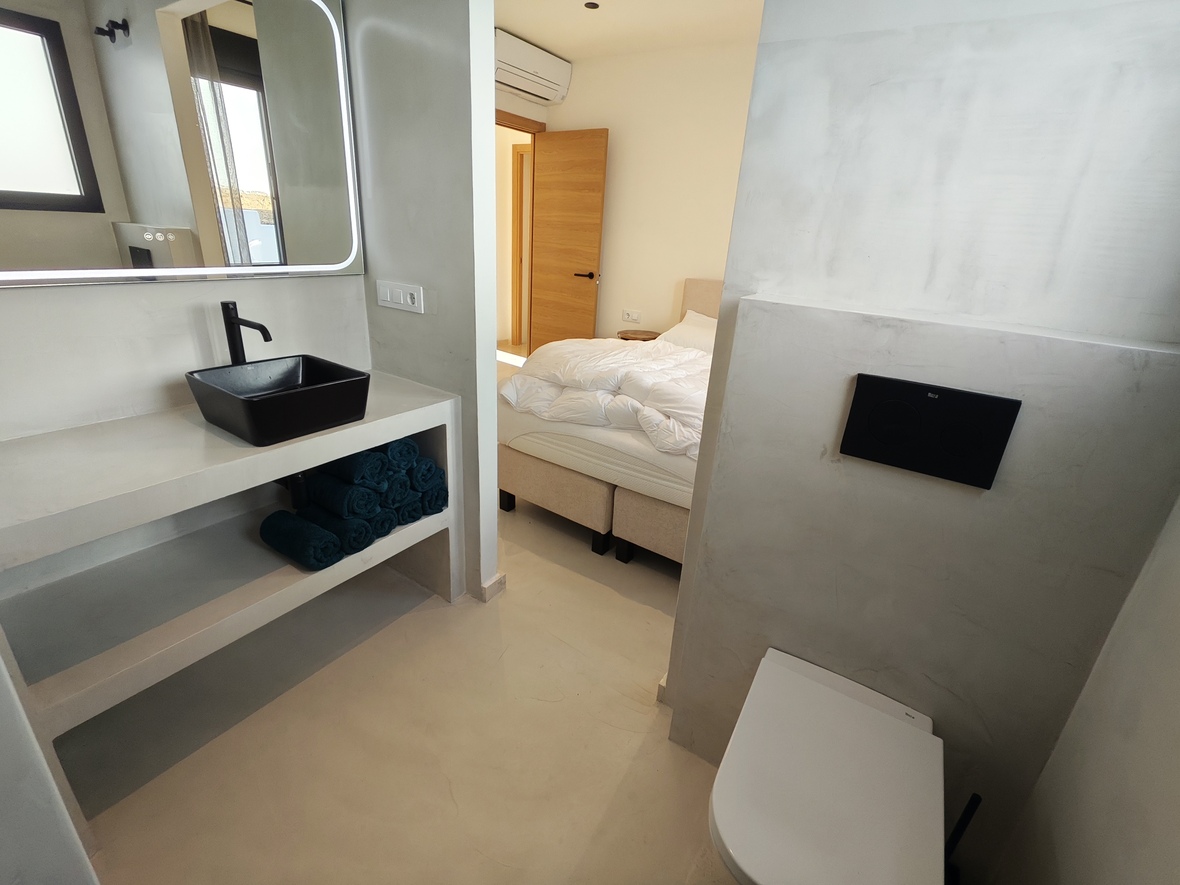 Salle de bains attenante moderne avec finitions en microciment, vasque noire et miroir rétroéclairé, offrant une vue sur la chambre à coucher.