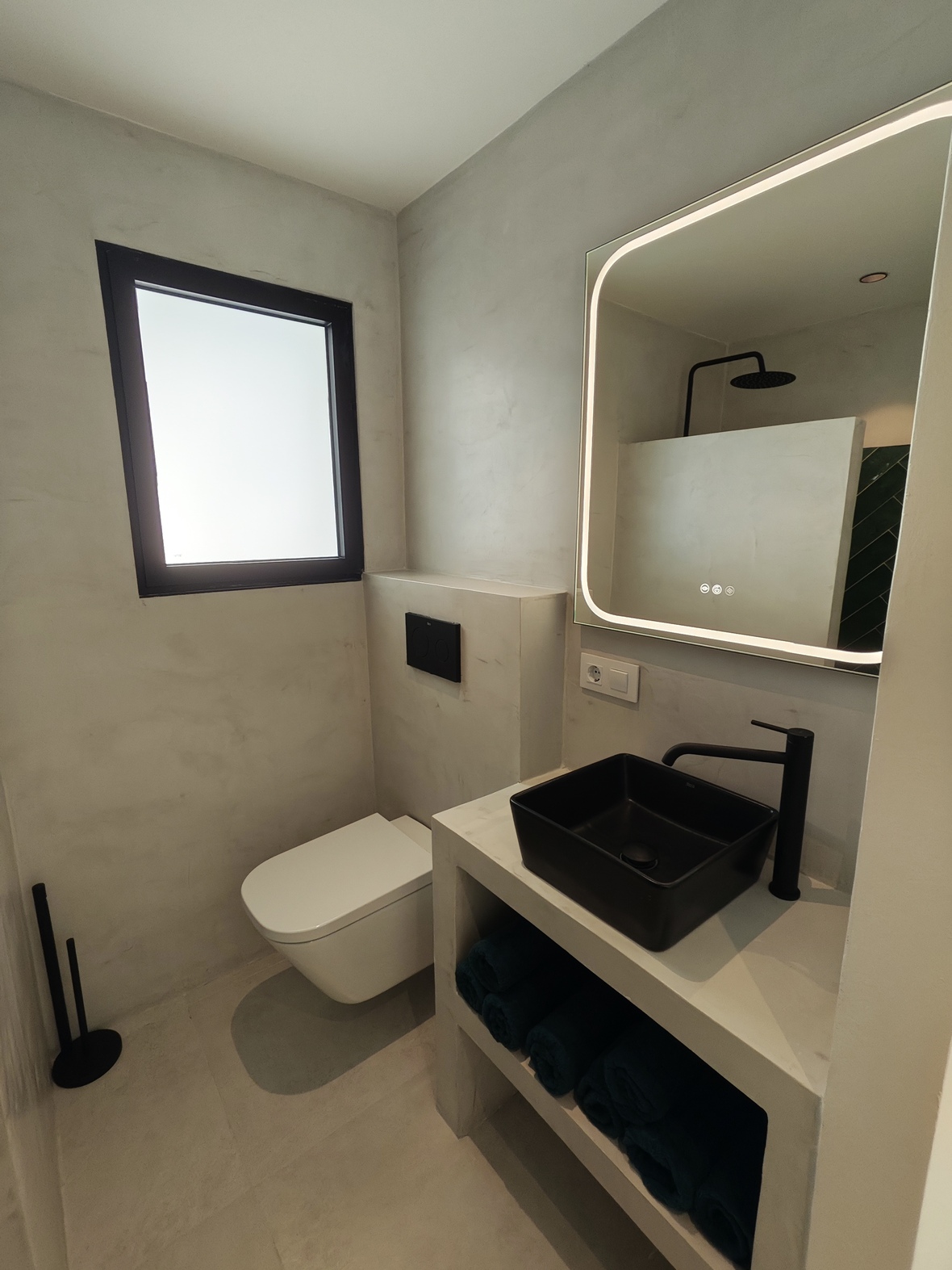 Salle d'eau en microciment avec vasque noire mate, miroir rétroéclairé et robinetterie noire. L'espace comprend un WC suspendu et des finitions minimalistes.